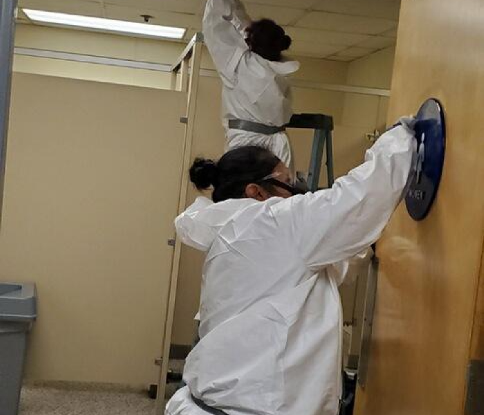 Two female technicians in Tyvek suits clean a commercial restroom.